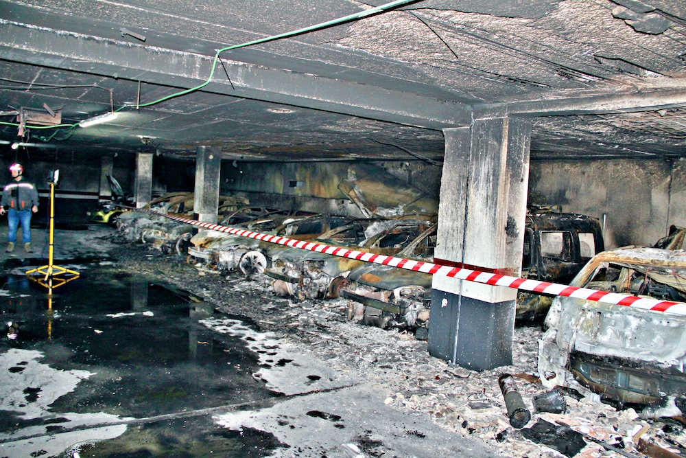 Vista general de aparcamiento subterráneo con múltiples vehículos destruidos por incendio, cinta de precaución perimetral y equipo de escáner láser durante la inspección forense