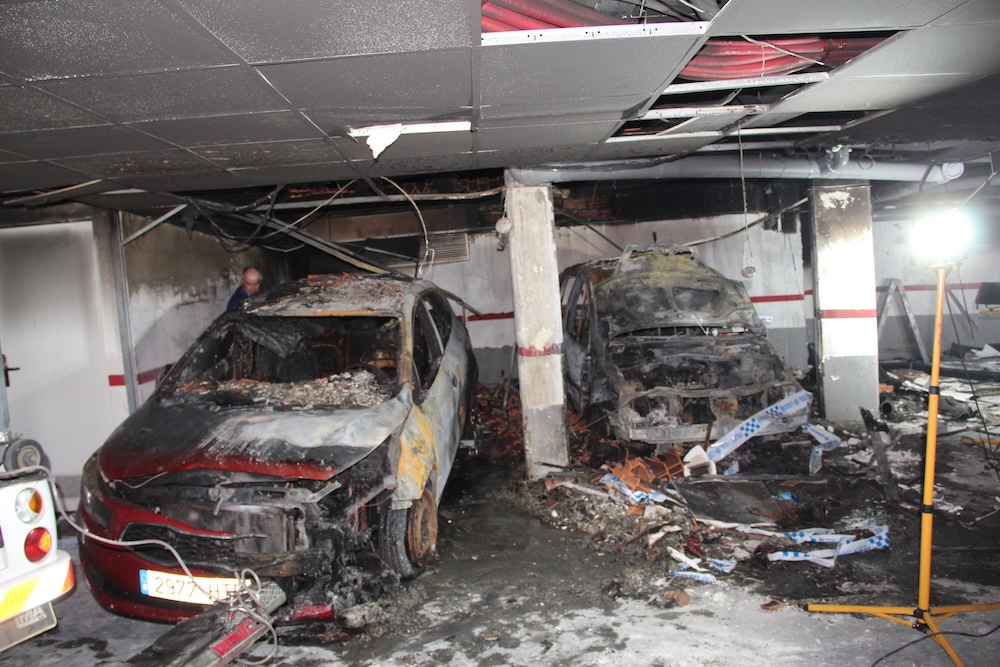 Investigador forense examinando dos vehículos calcinados en garaje comunitario tras incendio, con daños visibles en falso techo y señalización policial